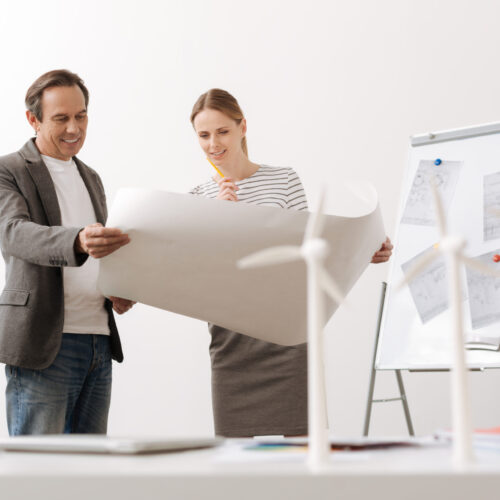 Work with positivity. Cheerful professioanl engineers holding a blueprint and discussign project while eco turbines models standing on the table